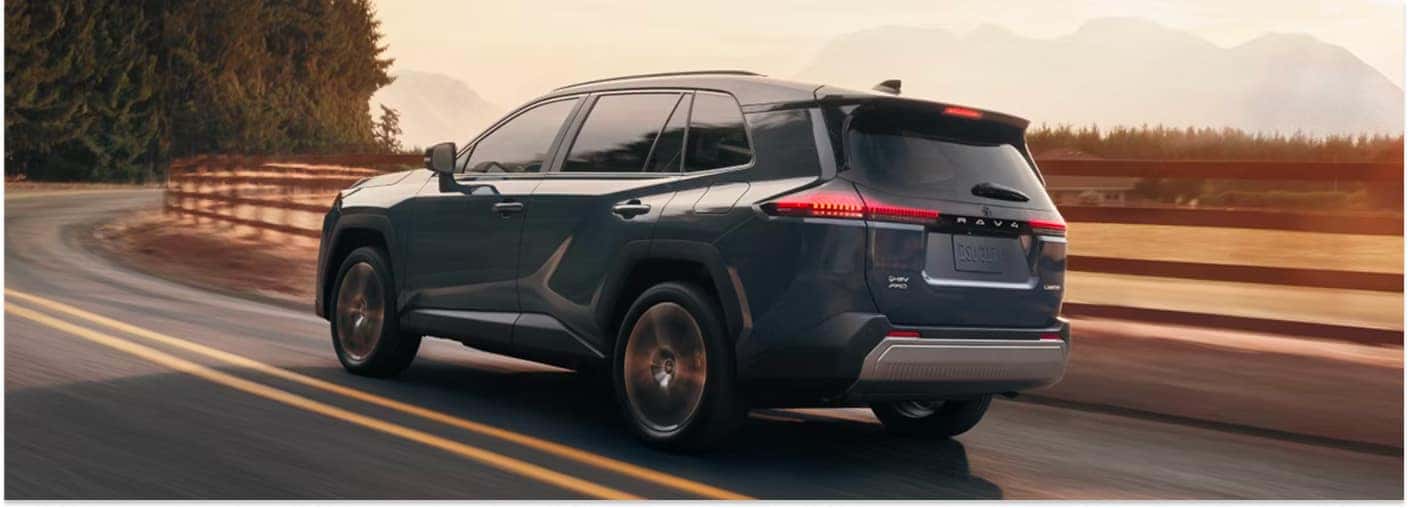 Rear view of a dark blue 2026 Toyota RAV4 driving on a two-lane road at sunset.