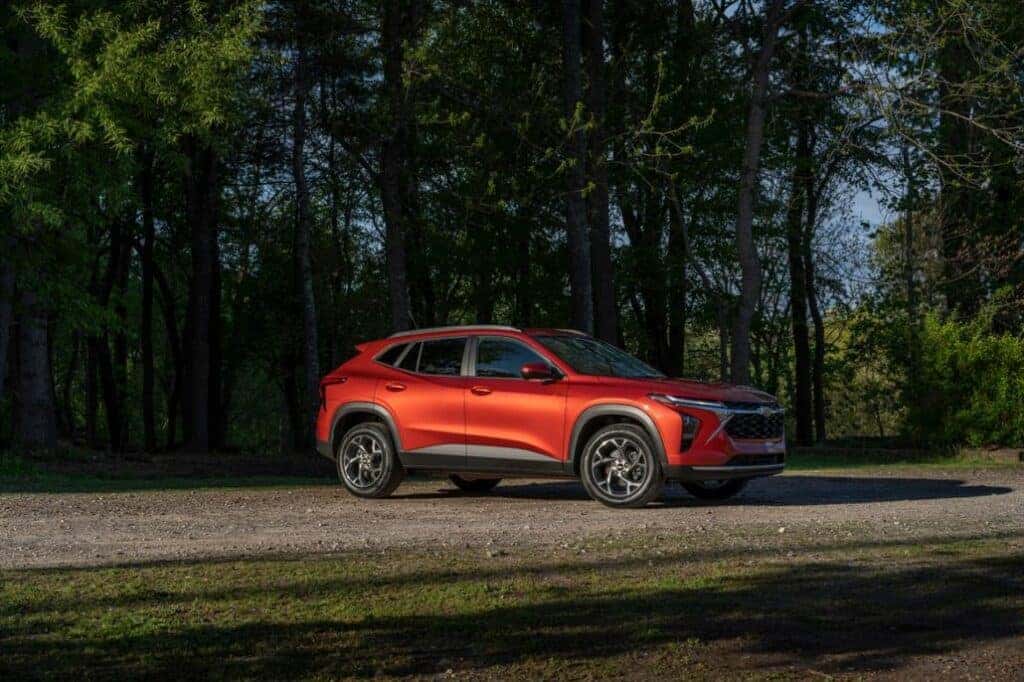 Passenger’s side 7/8 view of 2024 Chevrolet Trax LT in Cayenne Orange Metallic parked on a gravel road in front of trees.