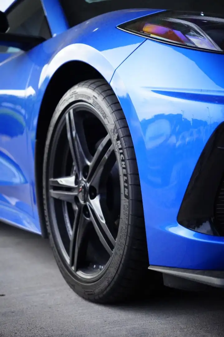 Close-up of the front wheel and blue body of a Chevrolet Corvette C8, highlighting a Michelin Pilot Sport All Season tire on a five-spoke dark alloy rim