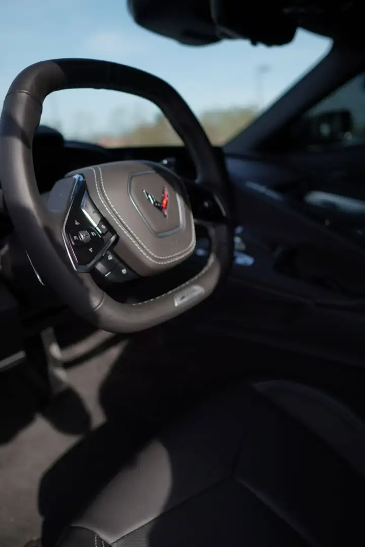 Interior cockpit view of a Chevrolet Corvette C8, showing a dark gray leather-wrapped squared steering wheel with contrast stitching, a centered Corvette crossed-flags logo badge, and integrated thumb controls