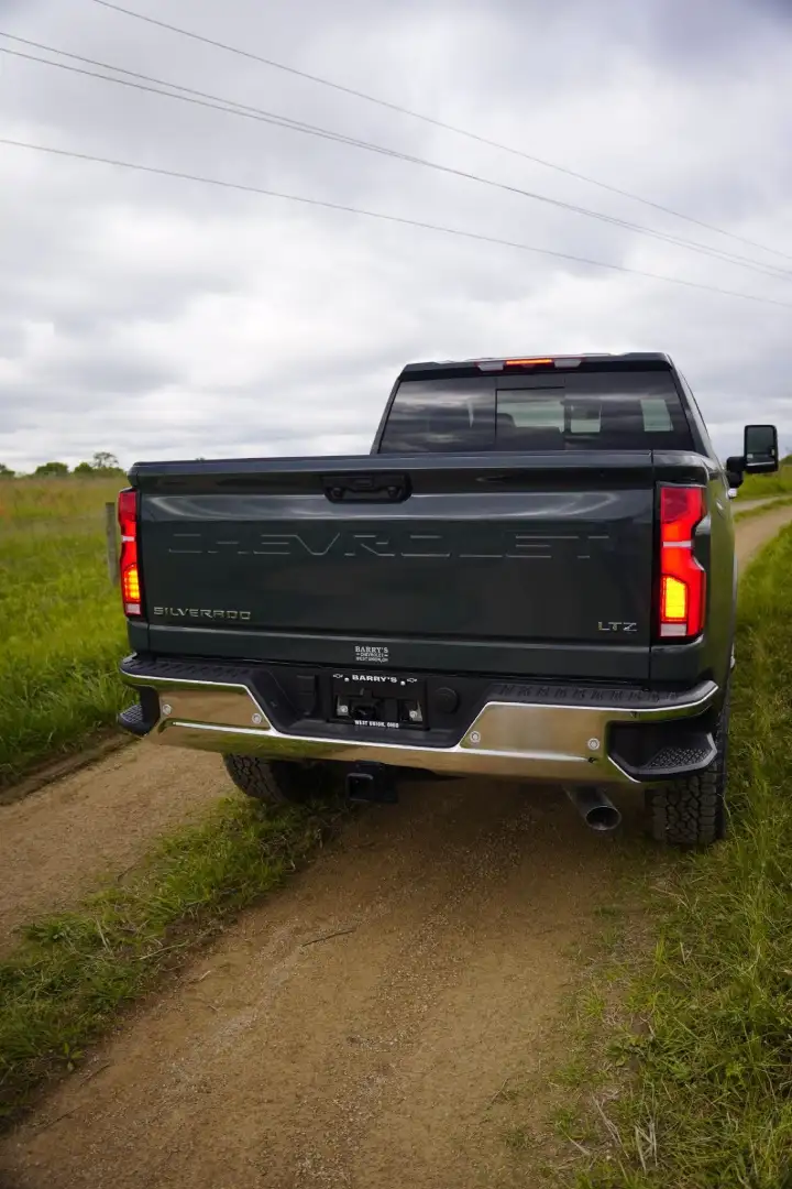 A rear view of a dark gray Chevrolet Silverado LTZ pickup truck parked on a dirt track through a grassy field under a cloudy sky.