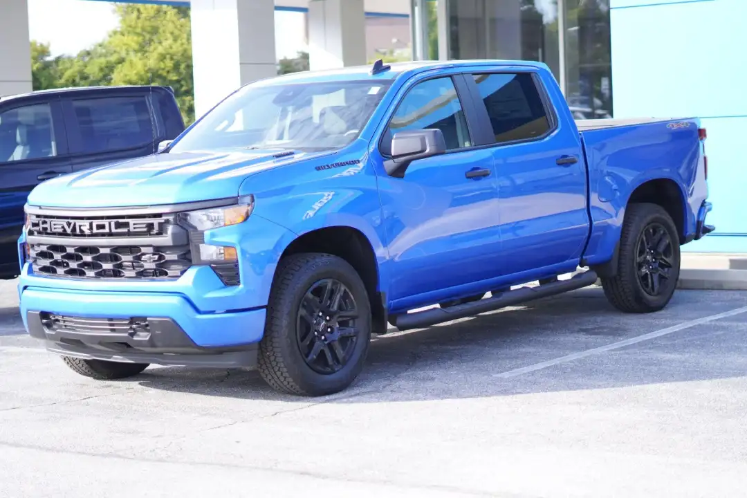A bright blue Chevrolet Silverado Custom crew cab pickup truck with black wheels and a black grille, parked on an asphalt lot in front of a dealership building.
