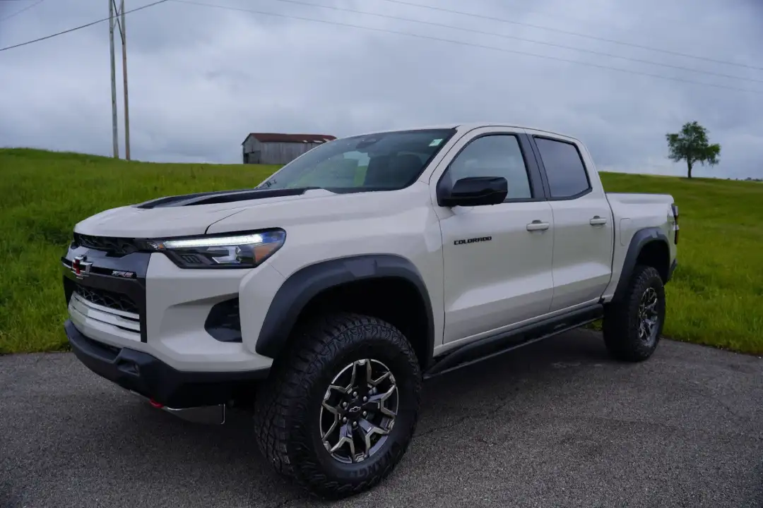 Front three-quarter view of a sand-colored or beige Chevrolet Colorado crew cab truck parked on asphalt with a green grassy hill and a rustic barn in the background under a cloudy sky