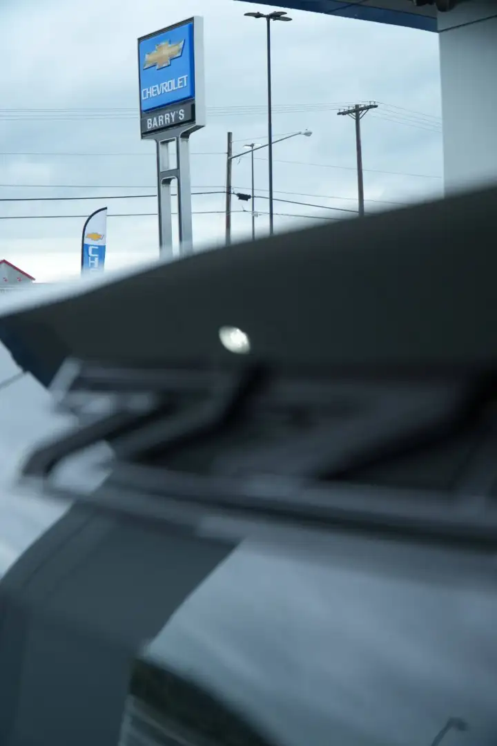 A low-angle view looking past a sharp, dark rear spoiler of a car toward a blue-and-white Barry's Chevrolet dealership sign against a cloudy sky.