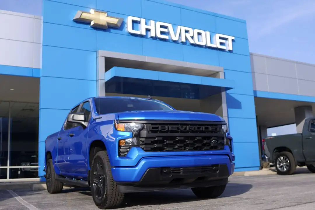 A bright blue Chevrolet Silverado Custom truck parked on the dealership lot directly beneath a modern blue-paneled Chevrolet dealership entrance sign