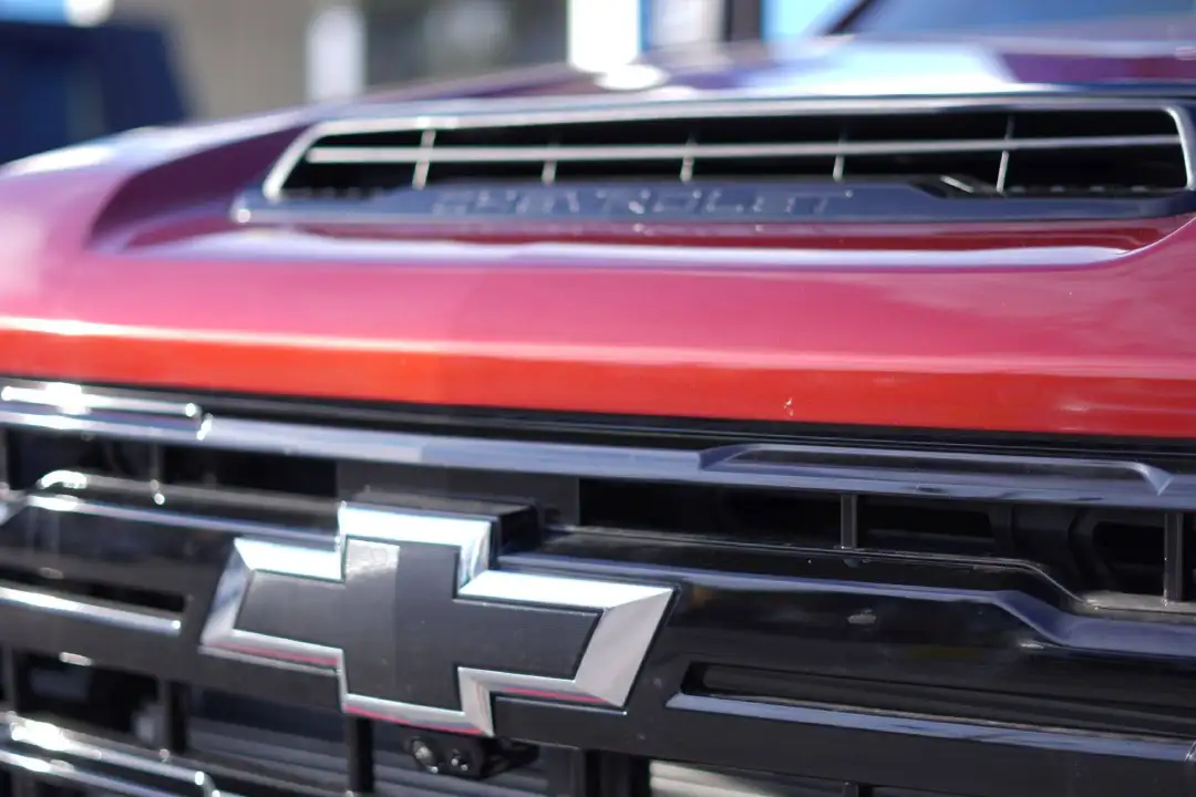 A close-up view focusing on a cherry red Chevrolet truck hood scoop with "CHEVROLET" lettering embossed on a black trim insert, situated above a glossy black grille and chrome bowtie emblem.