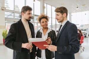 A salesperson talking to two others in a car dealership pointing to paperwork
