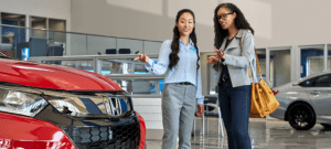 A woman car shopping pointing at red car with a salesperson in a dealership