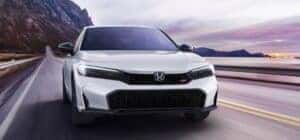 A white 2025 Honda Civic Si driving fast by a mountain, body of water, and sunset