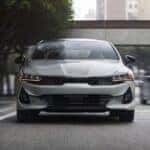 A gray 2024 Kia K5 driving front-facing driving on the road in the city