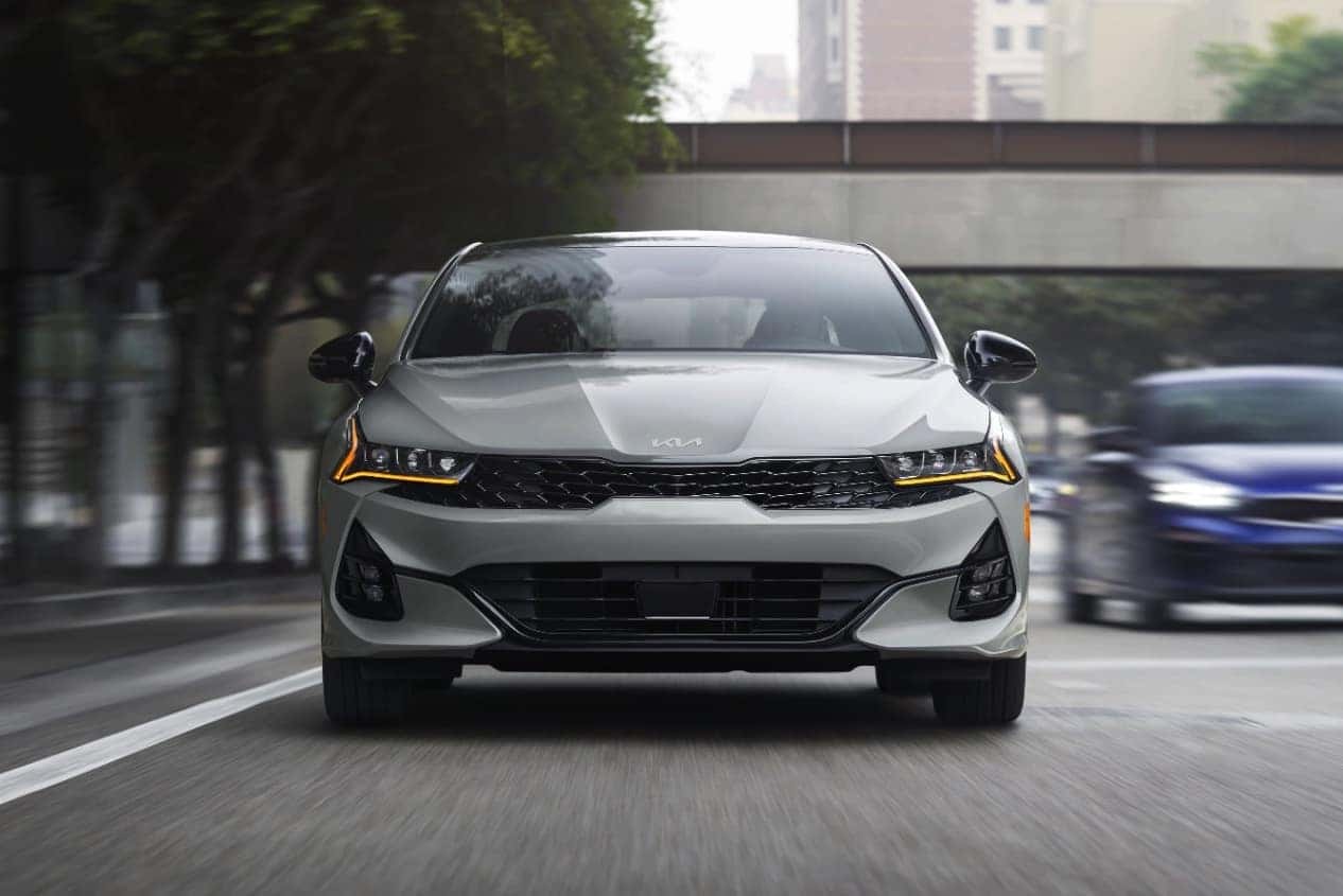 A gray 2024 Kia K5 driving front-facing driving on the road in the city
