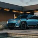 A blue 2026 Kia EV9 parked outside of a modern lit-up home