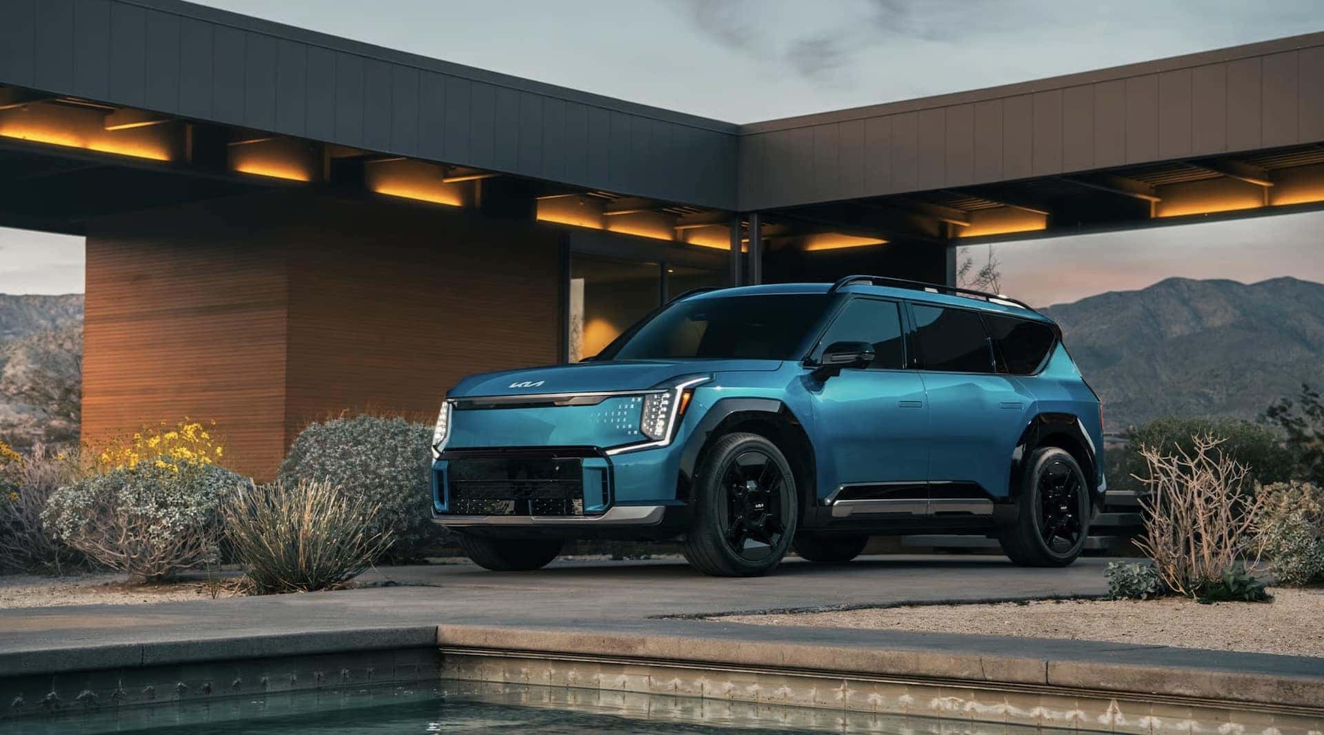 A blue 2026 Kia EV9 parked outside of a modern lit-up home