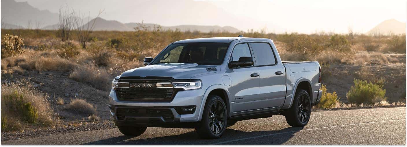 A front three-quarter view of a silver 2026 RAM 1500 pickup truck parked on a paved road in a desert landscape with mountains in the background.