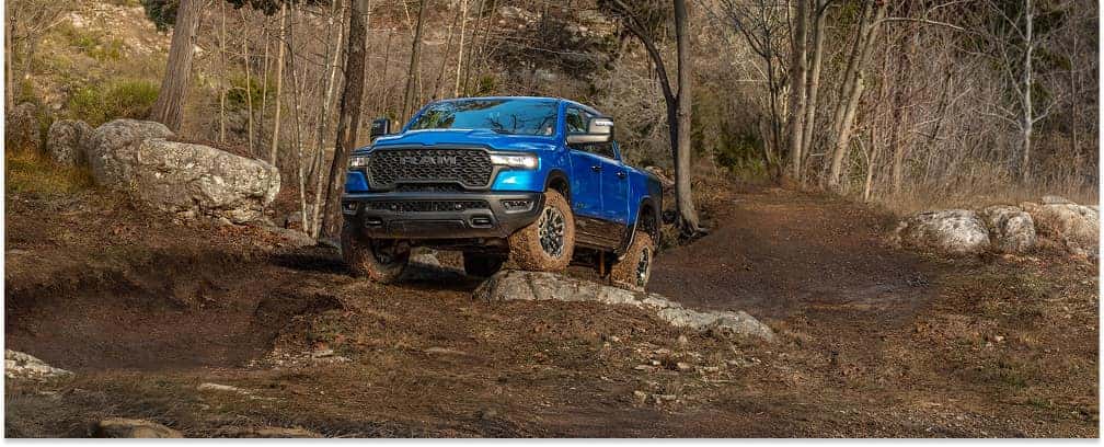 Front view of a blue 2026 RAM 1500 pickup truck driving on a rocky off-road trail through the woods.