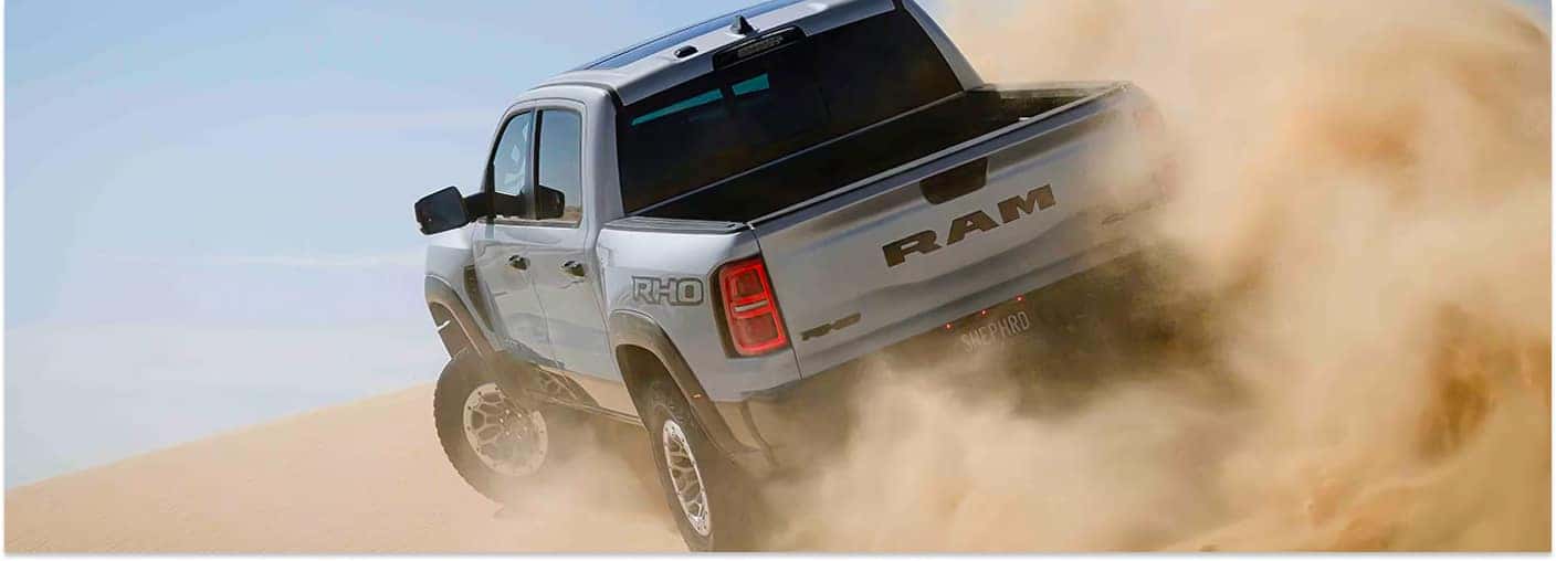 Silver 2026 RAM 1500 RHO pickup truck driving through sand dunes and kicking up desert dust.