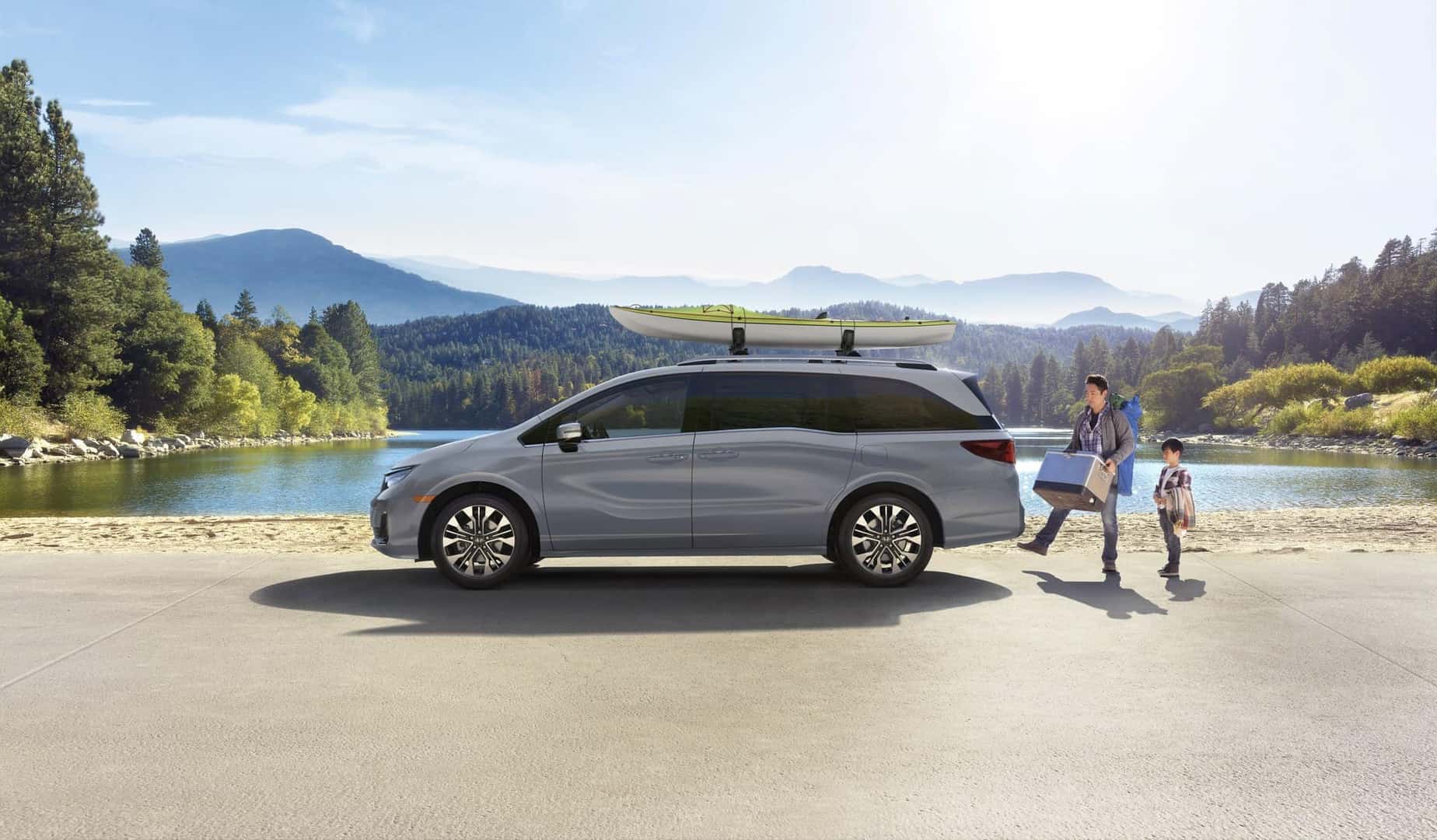 Gray Honda Odyssey with a roof-mounted kayak parked beside a lake as a parent and child unload gear