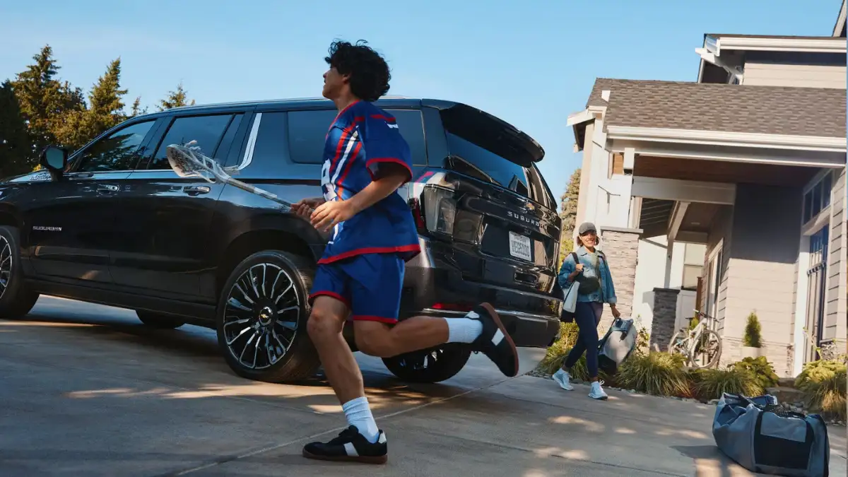 A black Chevrolet Suburban in a driveway with a kid with a lacross stick running in front of it