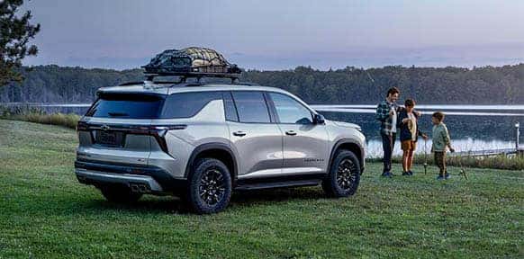 Silver Chevrolet SUV parked on a grassy field near a lake as a family prepares for an outdoor trip, highlighting the vehicle’s spaciousness and adventure-ready capability