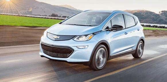 Light blue Chevrolet Bolt EV driving along a scenic country road at sunset, showcasing its modern electric design and efficiency