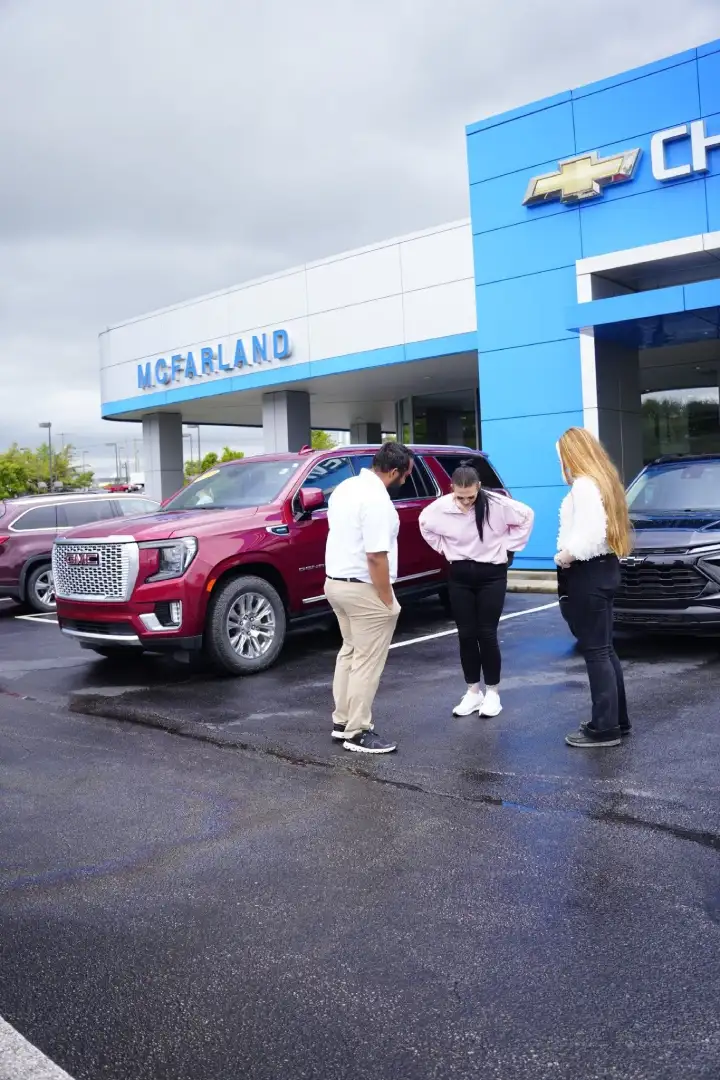 A salesperson talking with two customers outside the main entrance of McFarland Chevrolet dealership, parked next to a maroon GMC Yukon SUV and a black Chevrolet crossover on a damp lot