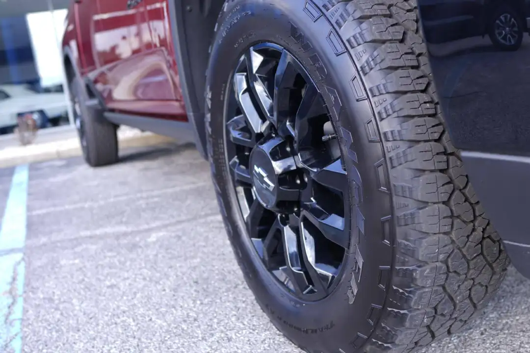 Close-up view of a rugged off-road tire on a glossy black wheel with a black Chevrolet bowtie center cap, mounted on a red truck parked on the dealership lot