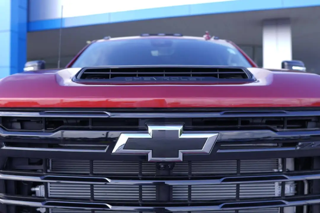 Close-up front view of a maroon Chevrolet Silverado HD truck, focusing on the blacked-out grille mesh, the prominent chrome bowtie emblem, and the functional hood scoop with integrated "CHEVROLET" lettering