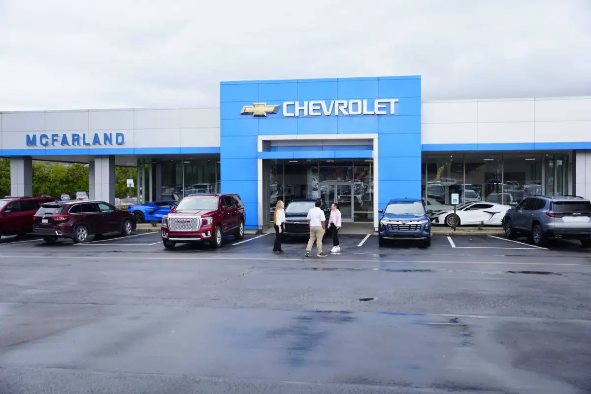 Salesperson greeting a couple outside the main entrance of McFarland Chevrolet dealership, with a blue Chevrolet Corvette, a red GMC Yukon, and other inventory parked in the front lot under an overcast sky