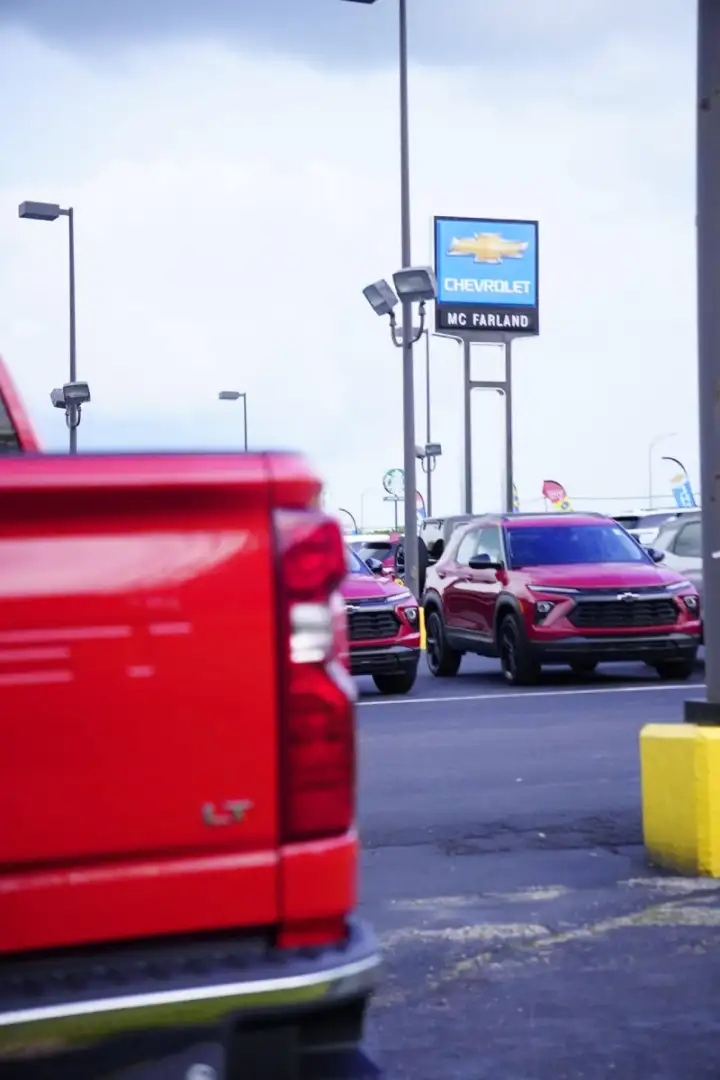 View across the dealership lot at McFarland Chevrolet, showing a red Chevrolet crossover parked in front of the blue "CHEVROLET MC FARLAND" sign, framed by the rear taillight of a red Silverado LT truck in the foreground