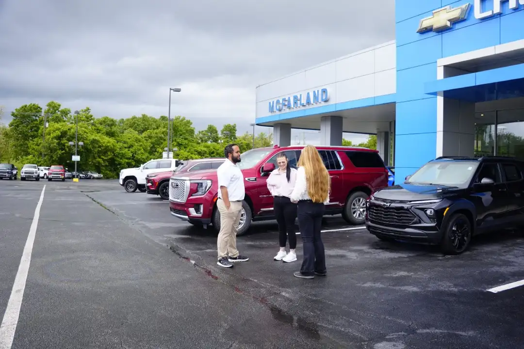 A McFarland Chevrolet salesperson talking with two customers on the dealership lot outside the main building entrance, next to a black Chevrolet crossover and a red GMC SUV