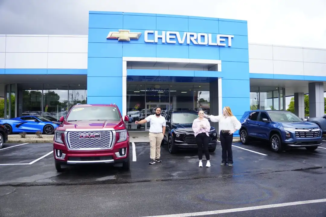 Customers standing with a salesperson outside the McFarland Chevrolet dealership entrance, celebrating a vehicle purchase next to a red GMC SUV and a black Chevrolet crossover