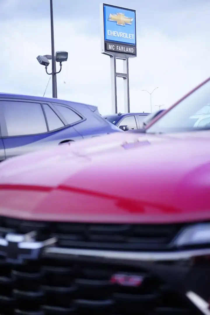 Close-up of a red Chevrolet vehicle parked on a dealership lot, with other vehicles and a tall blue "Chevrolet McFarland" sign visible in the background under an overcast sky