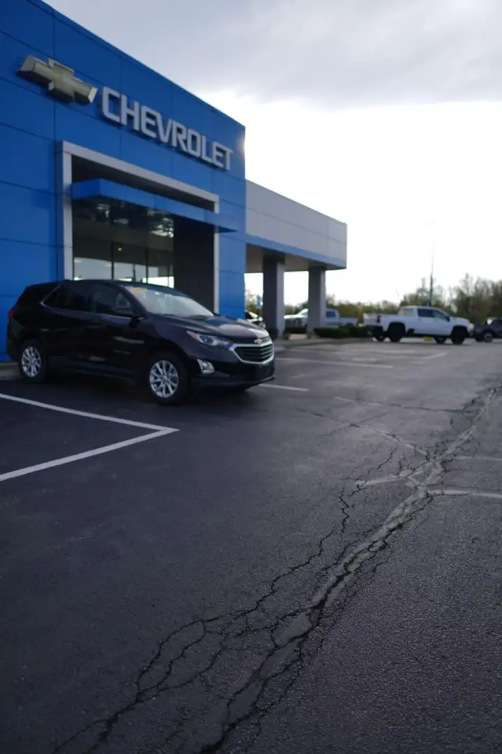 A dark gray Chevrolet SUV parked in front of the blue exterior of a Chevrolet dealership building, with an asphalt parking lot extending into the background