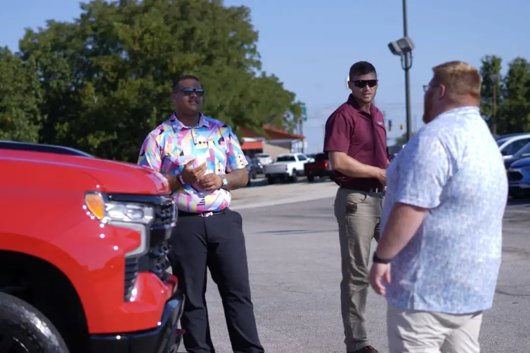 A car salesperson in a maroon polo shirt talking with two customers on an outdoor dealership lot next to a bright red truck.