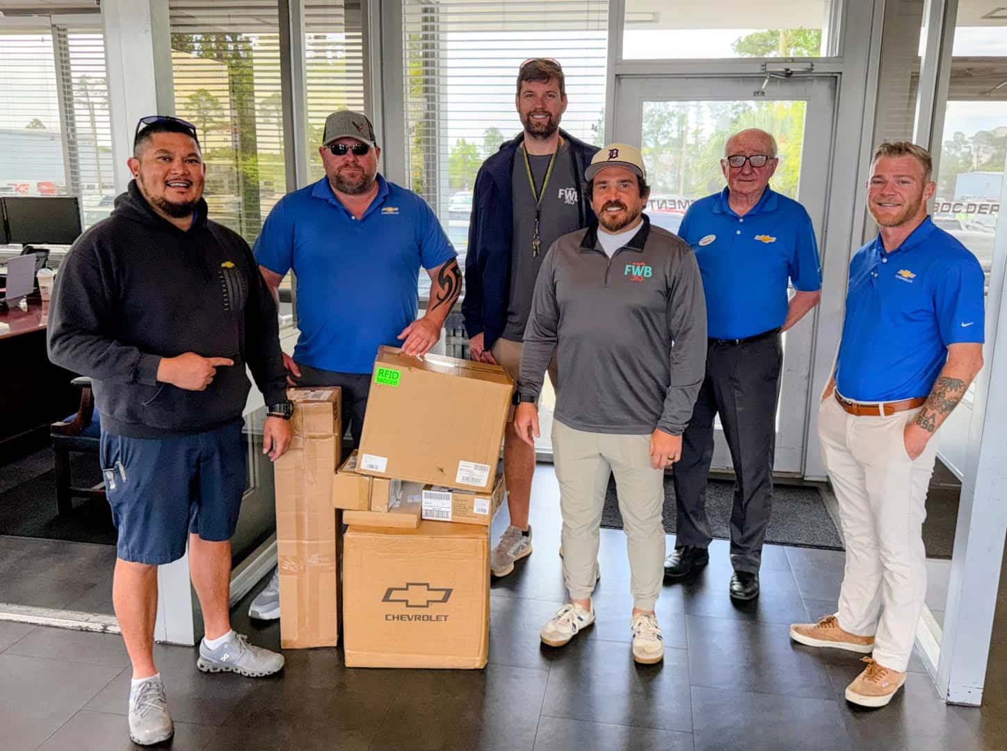 An image displaying six people standing around a pile of boxes of sports equipment smiling at the camera standing in the used car department at Preston Hood Chevrolet