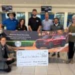 An image displaying Preston Hood Chevrolet employees presenting a check to the City of Fort Walton Beach inside the new car showroom next to two cars.
