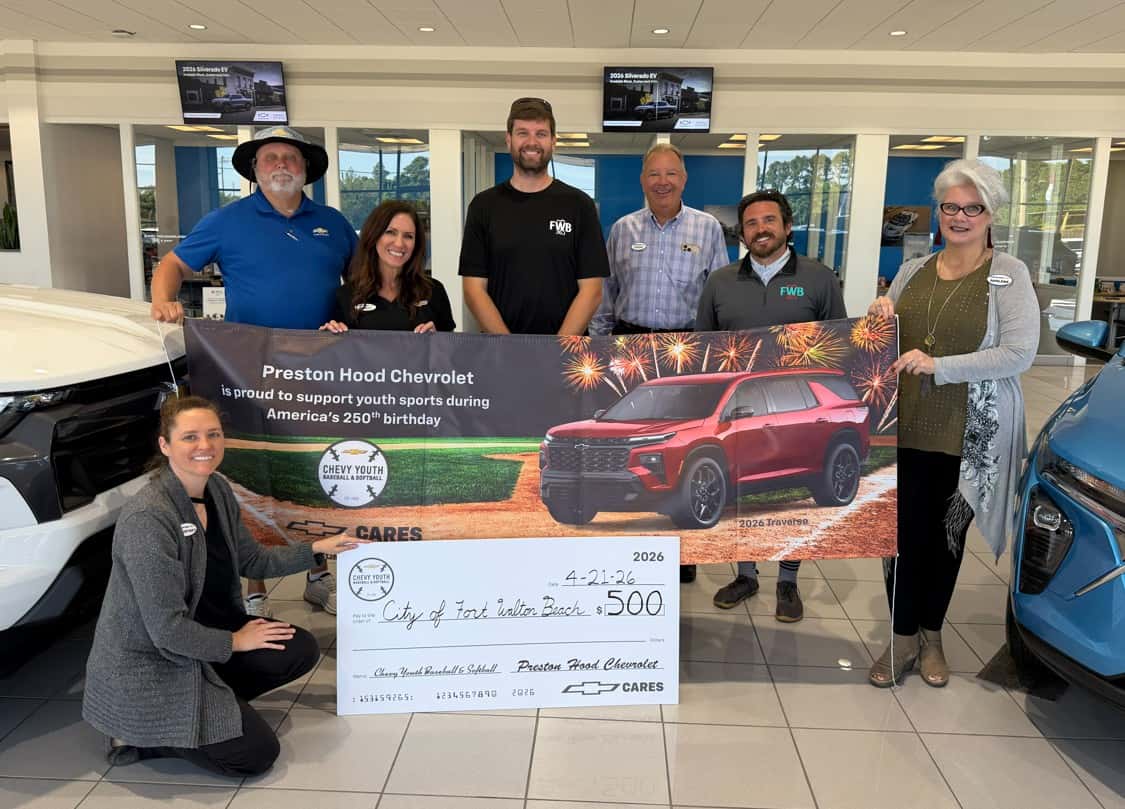 An image displaying Preston Hood Chevrolet employees presenting a check to the City of Fort Walton Beach inside the new car showroom next to two cars.