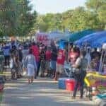 An image displaying a wide sidewalk with some pop-up tents lining the right side and palm trees on the left and a very large crowd of people going around the tents and partaking in the festival on a sunny afternoon