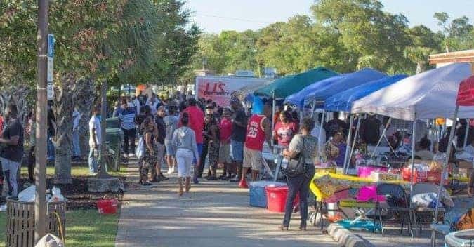 An image displaying a wide sidewalk with some pop-up tents lining the right side and palm trees on the left and a very large crowd of people going around the tents and partaking in the festival on a sunny afternoon