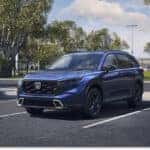The 2026-honda-cr-v-overview-blog-post.jpg image showcases a front three-quarter view of a blue 2026 Honda CR-V parked in an outdoor parking lot with trees and a sports field in the background.