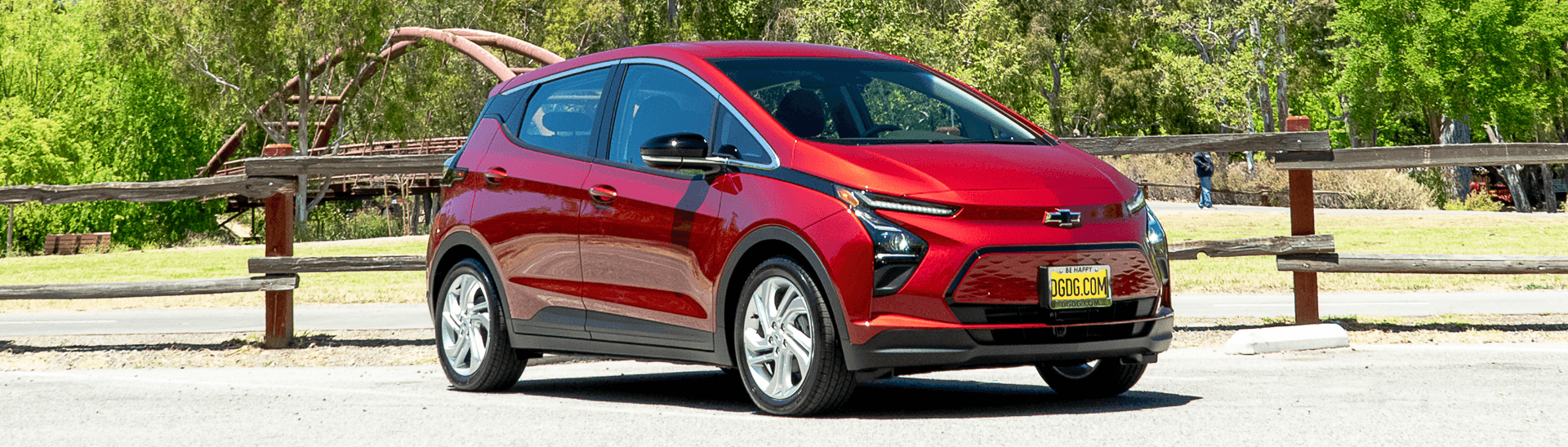 2022 Chevy Bolt EV 1LT in the color Cherry Red Tintcoat parked in a lot with a small bridge in the background.
